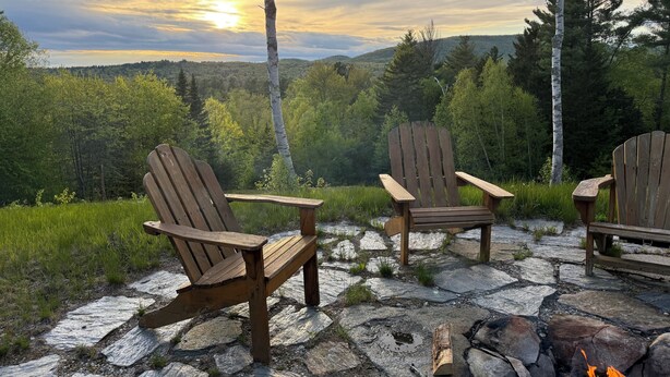Kaksi Adirondack-tyylistä puutarhatuolia kivipäällysteisellä terassilla, jonka keskellä on nuotiopaikka. Tuoleja ympäröi metsäinen maisema, ja sieltä avautuu näkymä auringonlaskuun.