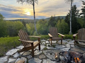 Two Adirondack chairs on a stone patio with a fire pit, surrounded by a forested landscape and a view of the sunset. - Cozy mountain cabin w/Mountain View, hot tub, game room, and adventure for all (Orange)