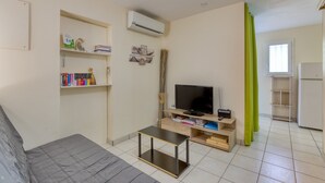 Ein kompakter Wohnraum mit einer Couch, einem kleinen Fernseher, einem Bücherregal und einem Fenster mit Vorhängen.