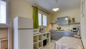 Eine kleine Küche mit Kühlschrank, Mikrowelle und Spüle. Ein Esstisch mit Stuhl und ein Fenster mit Vorhängen sind ebenfalls vorhanden.