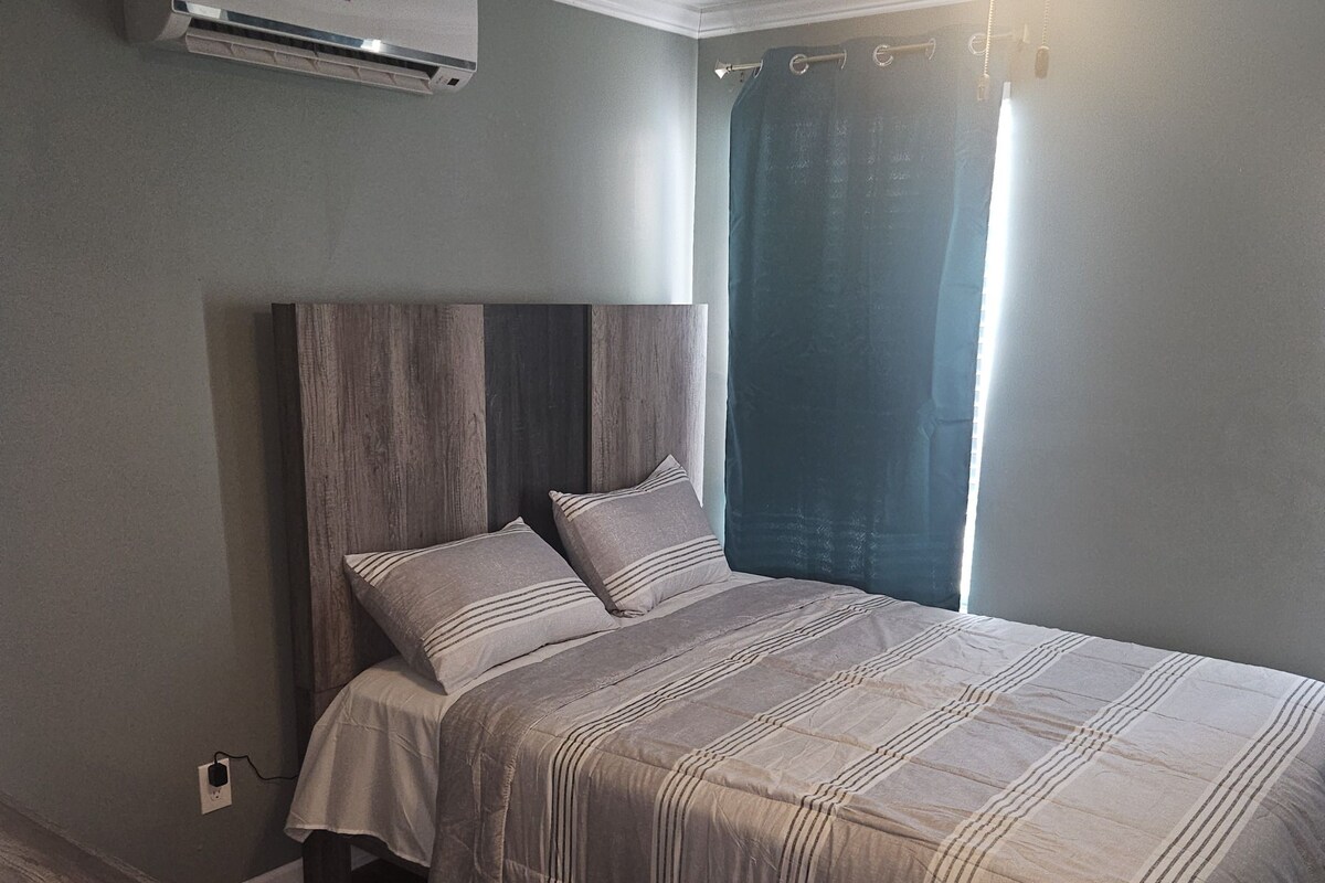 A bedroom with a bed, a ceiling fan, an air conditioning unit, and a window with curtains.