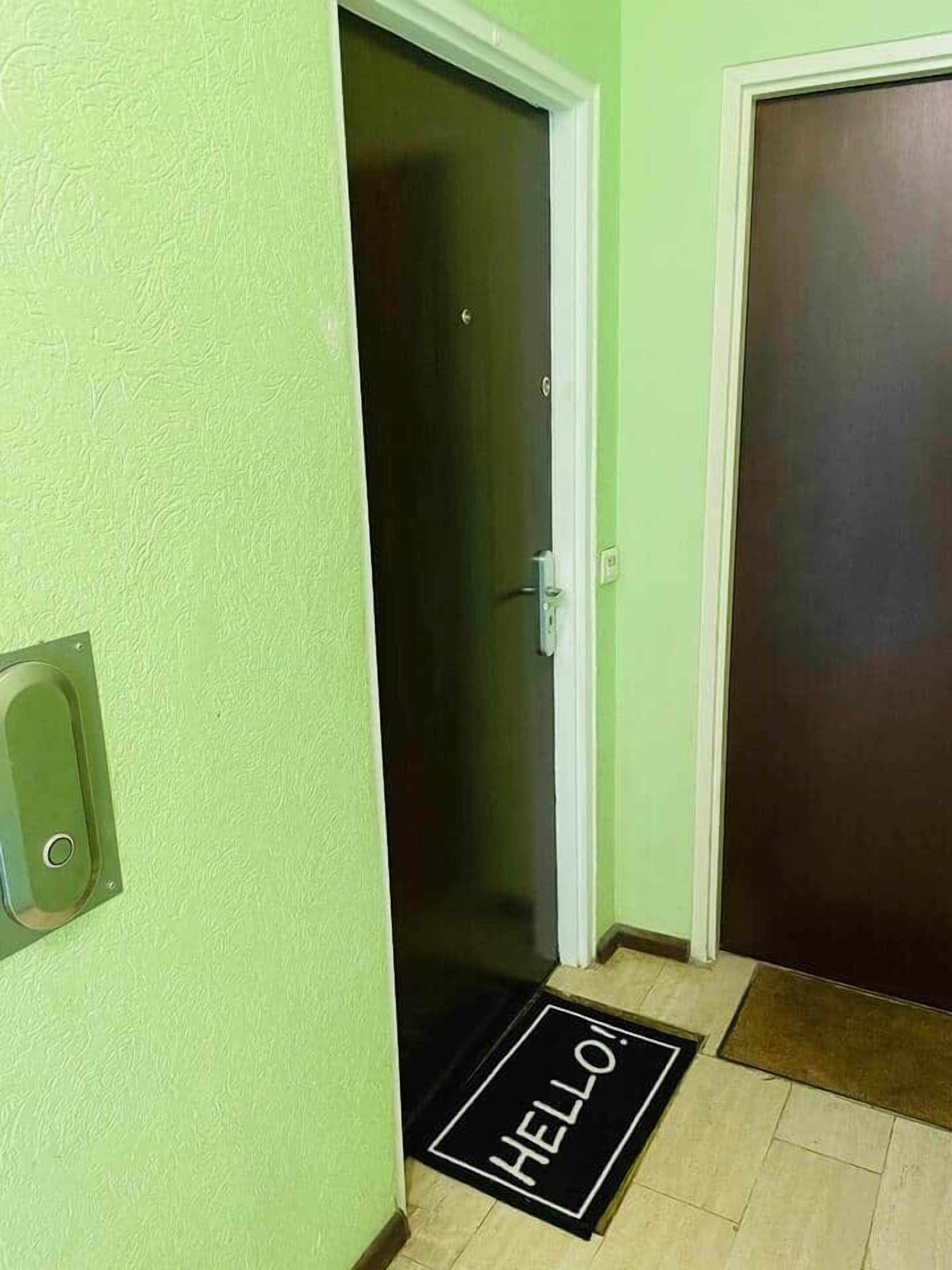 Ein Flur mit grüner Wand, einer geschlossenen Tür und einer schwarzen Fußmatte mit der Aufschrift "HELLO!".
