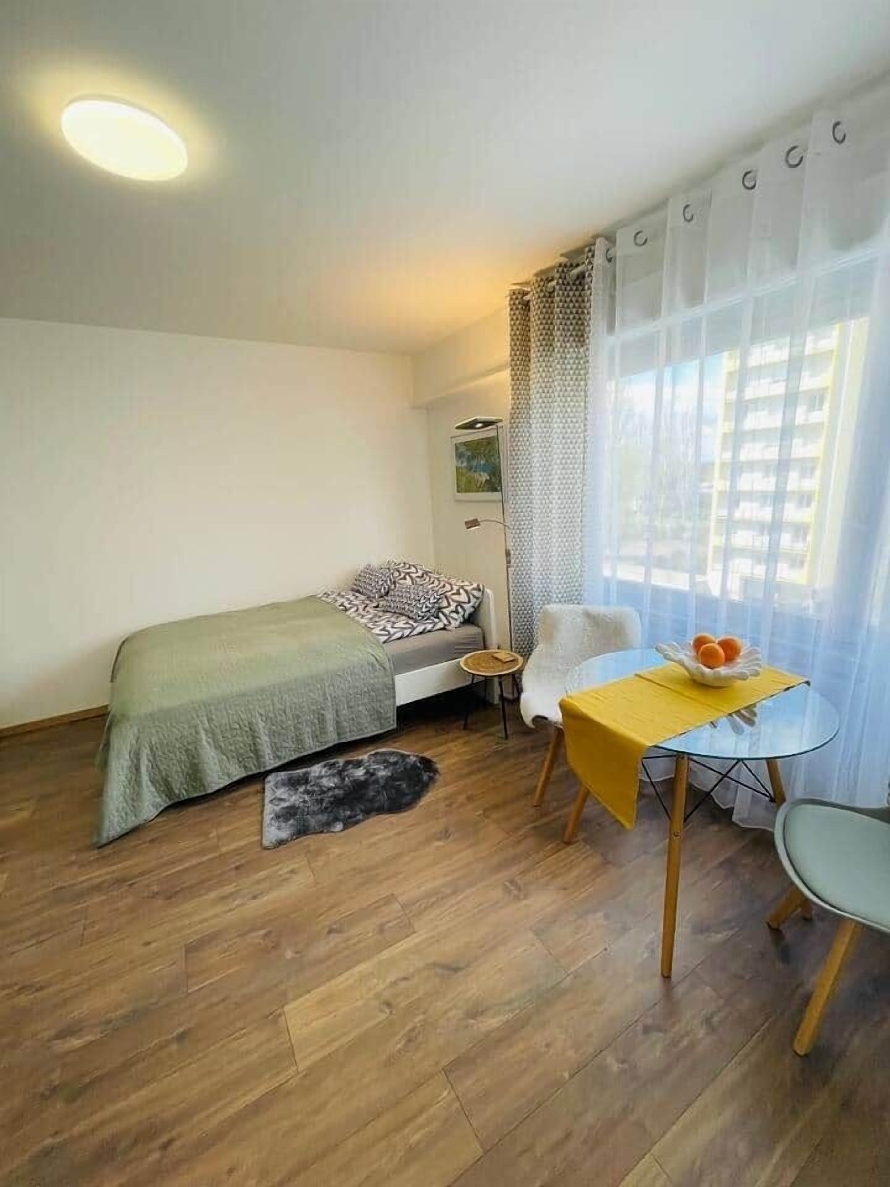 Una habitación pequeña y luminosa con una cama, una mesita con un frutero, una silla y una ventana con cortinas translúcidas.