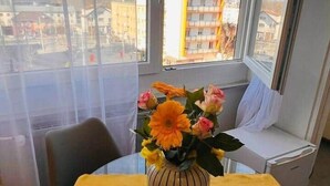 A room with a yellow table, a vase with flowers, and a view of a building through large windows.