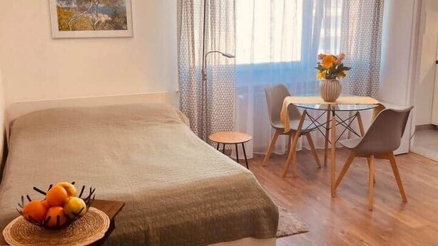 A bedroom with a bed, a small table with chairs, a vase with flowers, and a painting on the wall.