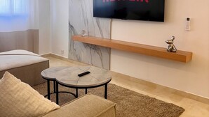 A modern living room with a flat-screen TV mounted on the wall, a wooden shelf below it, a round coffee table, and a beige sofa. - Stylish & Cozy | Downtown 2BR Netflix+Fast Wifi•AC (rabat)