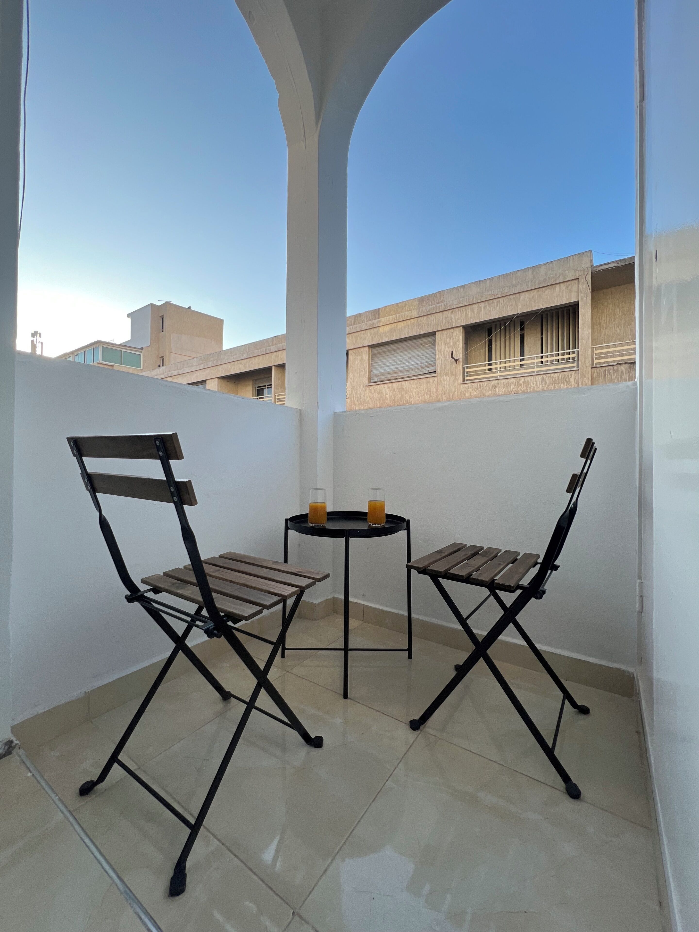 Varanda com duas cadeiras de madeira e uma pequena mesa redonda, vista desobstruída de edifícios ao fundo e piso de azulejos.