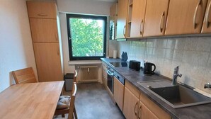 A modern kitchen with wooden cabinets, a built-in oven, a large window, and a dining area with a table and chairs. - Dora - Bergheim near Cologne (Bergheim)