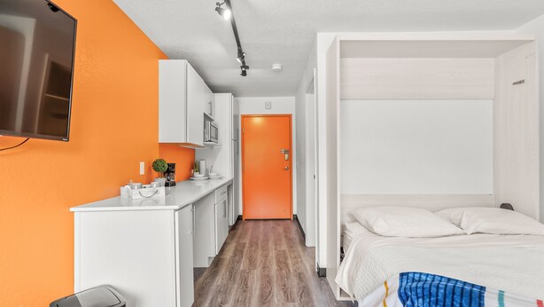 Ein kleines Hotelzimmer mit einer Küchenzeile, einem Bett mit bunter Decke und einer orangenen Tür.
