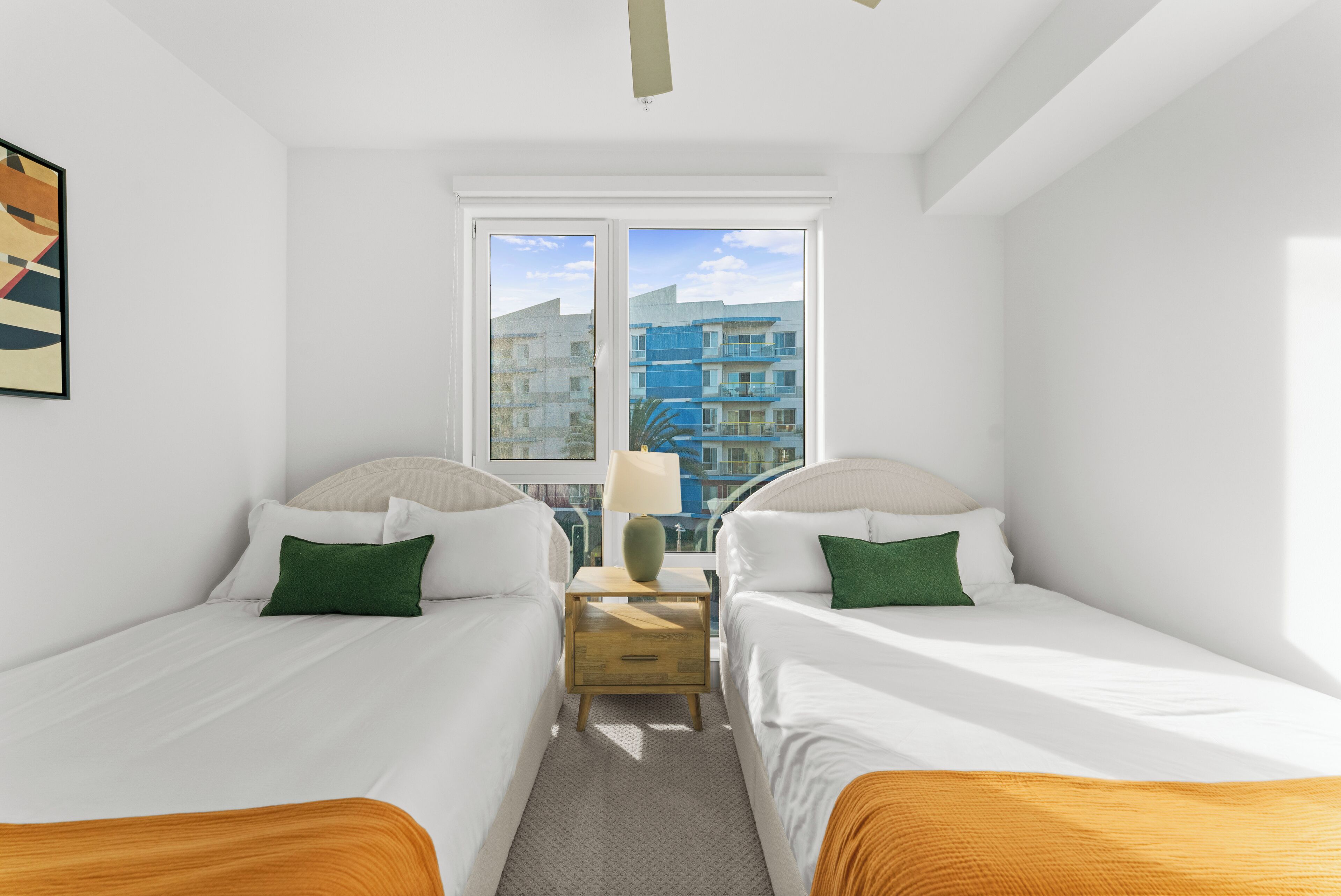 A hotel room with two beds, a window with a view of buildings, and a small table with a lamp.