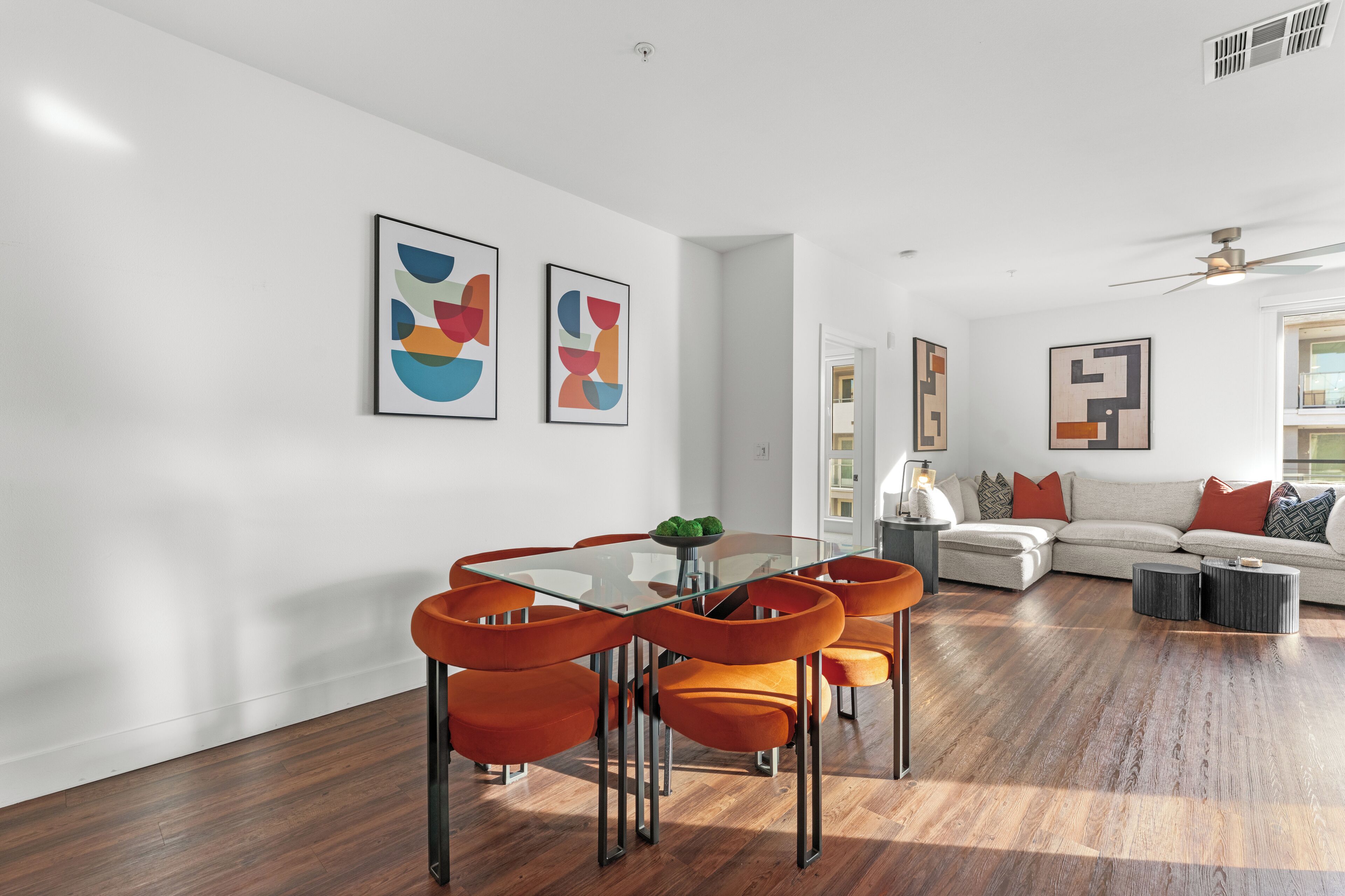 A modern living room with a glass dining table, orange chairs, a sofa, and abstract wall art.