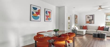 A modern living room with a glass dining table, orange chairs, a sofa, and abstract wall art.