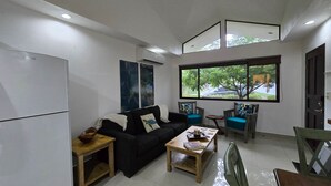 Un salon moderne avec un canapé noir, une table basse en bois et une petite table à manger. On trouve un réfrigérateur sur la gauche et une fenêtre donnant sur des arbres et une voiture à l’extérieur.