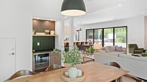 A modern kitchen with a dining area, featuring a large wooden table, a hanging pendant light, and a view of the outdoors through large windows. - Blackwood Shores - 6 bed, pool, hot tub, game room (Lakeside)