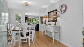 Un coin repas avec une table et des chaises blanches, une console blanche agrémentée de lampes, et une vue sur la verdure à l’extérieur.