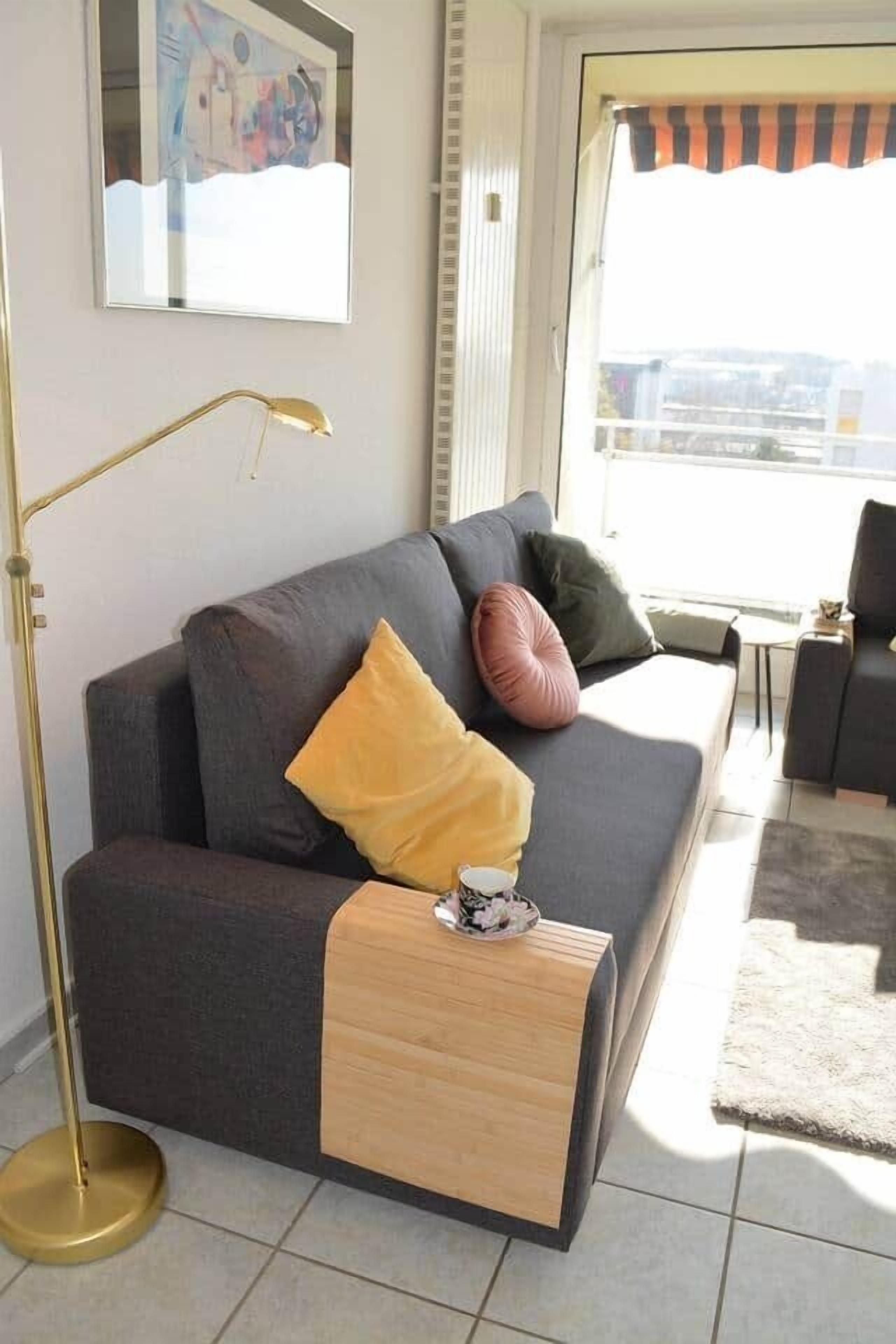 A modern living room with a grey sofa, a small wooden side table, a floor lamp, and a large window with a view.