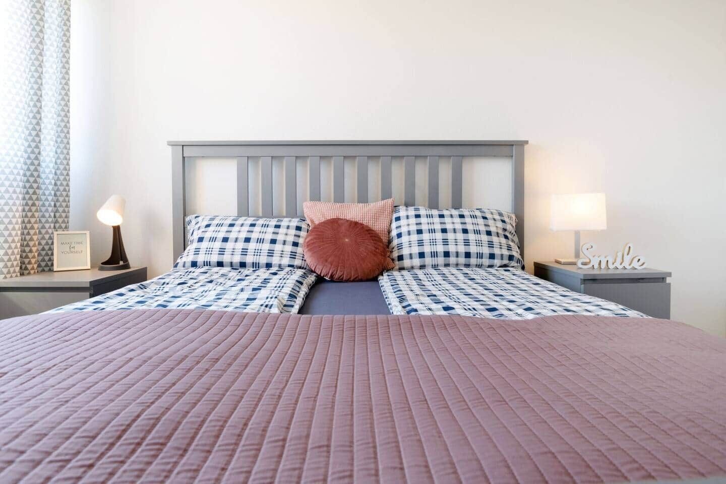 Cama bem arrumada com edredom xadrez e travesseiros, luminária de cabeceira e parede com padrão geométrico.