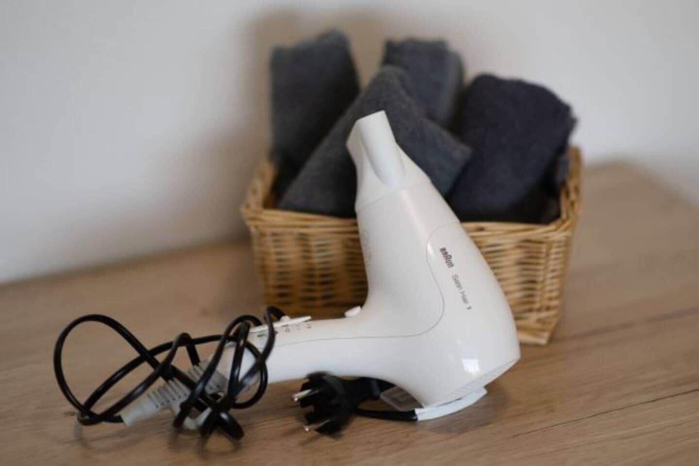 Un casc d'eixugar cabells blanc amb un cable d'alimentació sobre una superfície de fusta, al costat d'una cistella de vimet plena de tovalloles grises plegades.