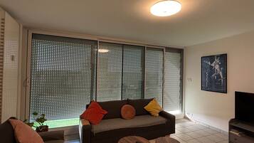 A living room with a sofa, two small tables, a TV, and a large window with blinds.