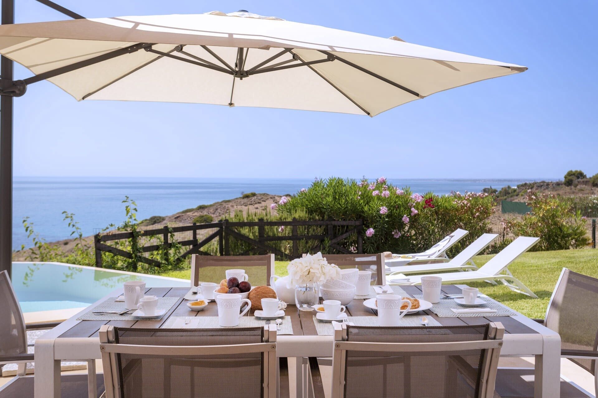 A dining table set for two with a view of the ocean, a wooden fence, and a pool in the background.