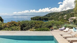 A pool area with lounge chairs, a view of the sea, and a hillside with buildings. - Josephine House - Four Bedroom Villa, Sleeps 8 (Kalami)