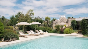 A pool with lounge chairs and umbrellas, surrounded by greenery and a stone building. - Trullo Terra Dolce - Four Bedroom Villa, Sleeps 8 (Ostuni)