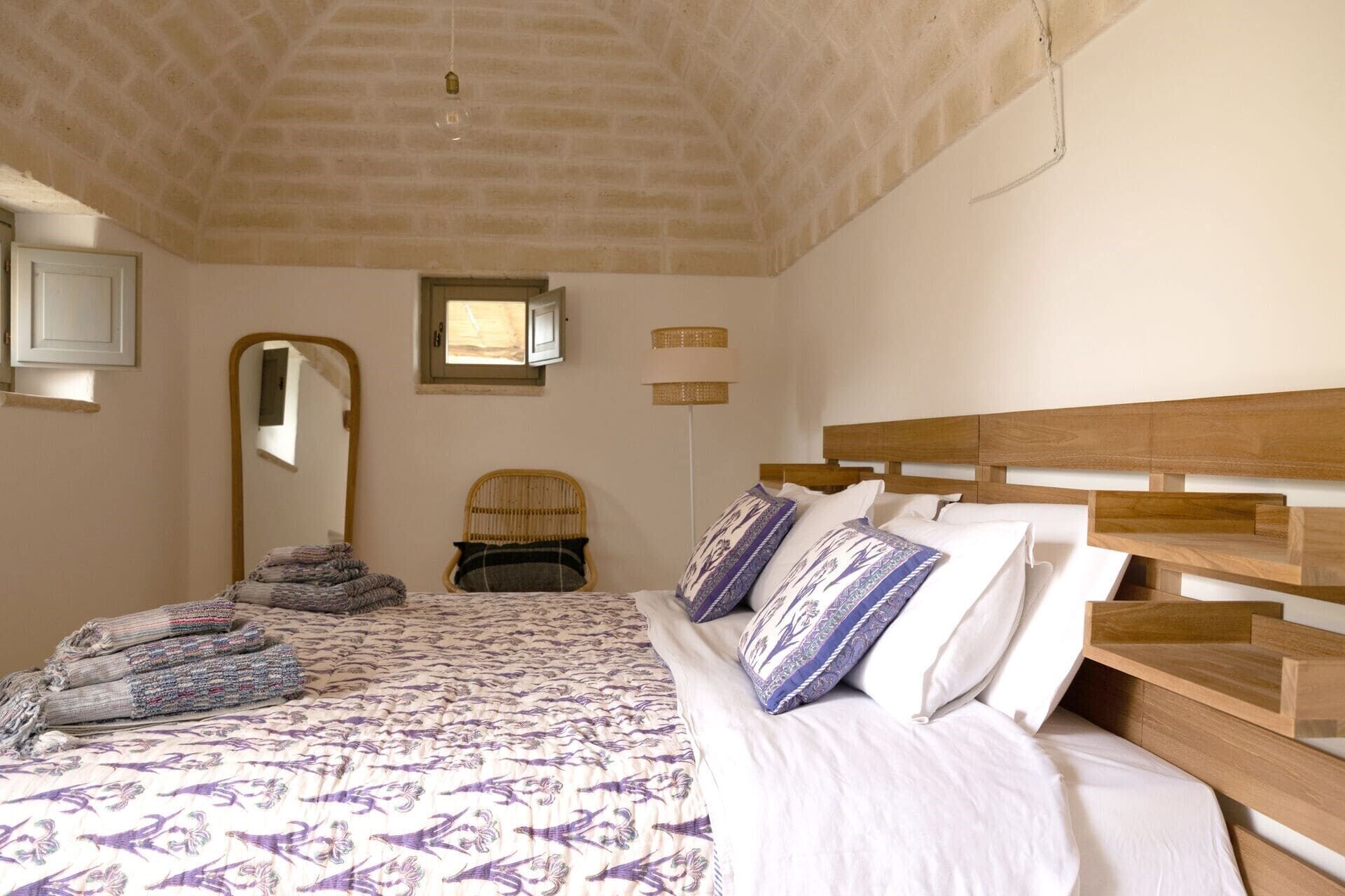 Un dormitorio con una cama, cabecera de madera, un espejo, una ventana y un estante fijado a la pared.