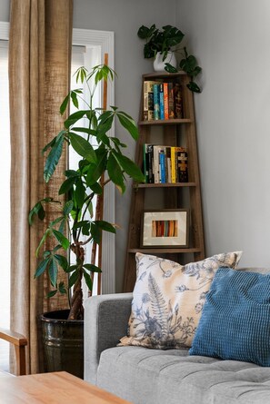 Ein Wohnzimmer mit grauem Sofa, einem blauen Kissen, einem Holzregal, einer Topfpflanze und einem Fenster mit Vorhängen.