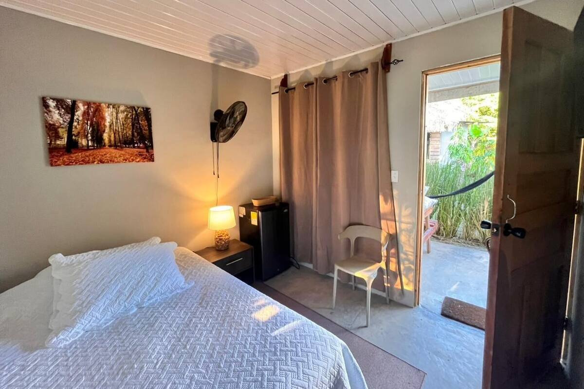 A bedroom with a bed, bedside lamp, a small table, a chair, a painting, and a door leading to an outdoor area.