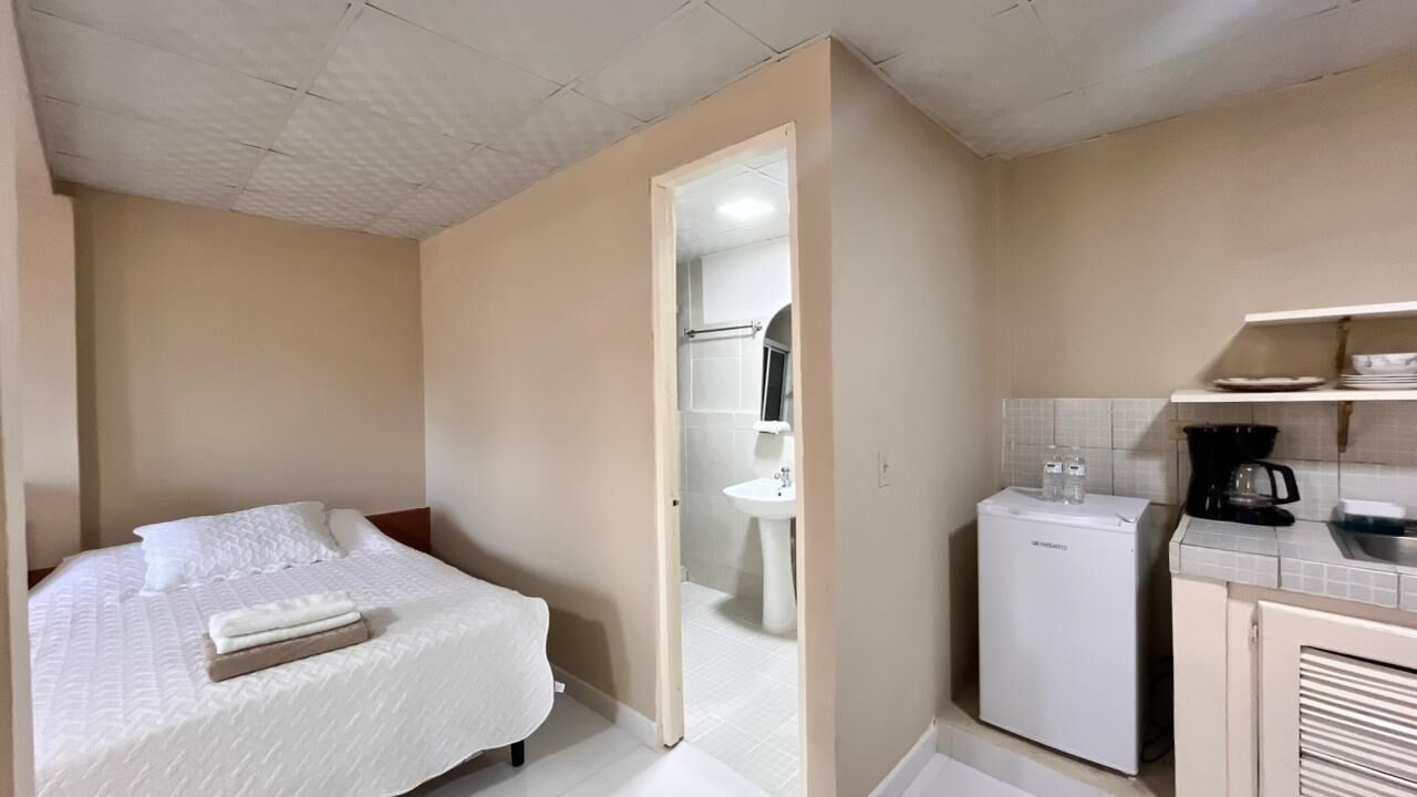 Habitación de hotel compacta con cama, una kitchenette pequeña y un baño visible a través de una puerta abierta.