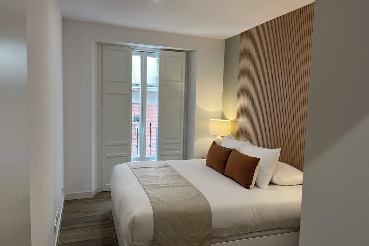 Una camera d'albergo con un letto, un comodino con una lampada, una parete rivestita in legno e una porta che dà su un balcone.