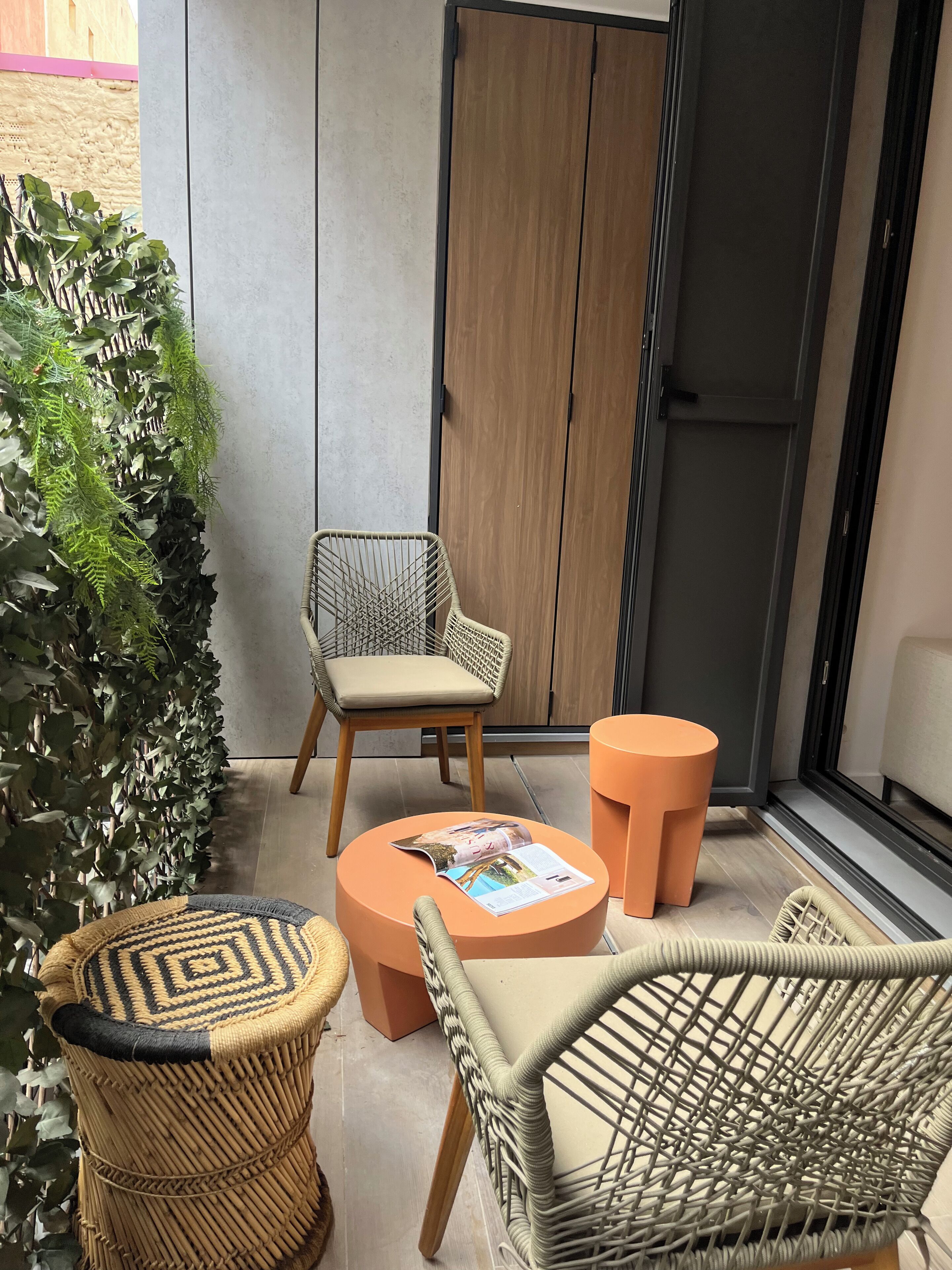 Un piccolo spazio esterno con sedute in vimini, tavolino rotondo, una porta in legno e una pianta sospesa al muro.
