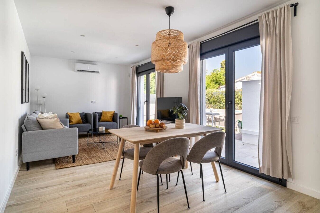 Una sala de estar moderna con comedor, un sofá gris, una mesa de comedor de madera y un amplio ventanal con cortinas.