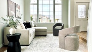 Ein Wohnzimmer mit einer Eckcouch, einem runden Couchtisch, einem grauen Sessel und einem großen Fenster mit Blick auf Häuser.
