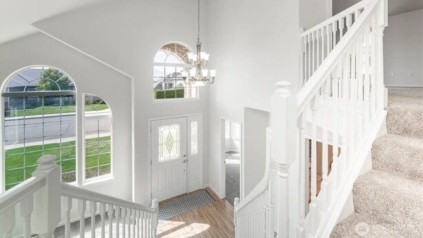 Ein geräumiger Innenraum mit hoher Decke, einer Treppe mit weißen Geländern und großen Fenstern, die Blick auf eine grüne Landschaft bieten.