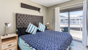 Une chambre avec un grand lit, une table de chevet en bois, une lampe de chevet et une vue sur l’extérieur grâce à une grande fenêtre.