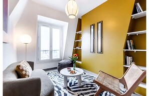 Ein modernes Wohnzimmer mit einer Couch, einem runden Couchtisch mit Blumenvase, einem gemusterten Teppich und einem Bücherregal an einer gelben Wand.