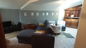 A living room with a sofa, ottoman, and a TV. There are framed pictures on the wall and a wooden cabinet in the background.