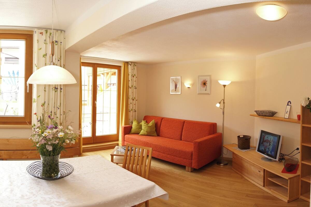 A cozy living room with a wooden bench, a table with a vase of flowers, a red sofa, and a TV on a wooden stand.