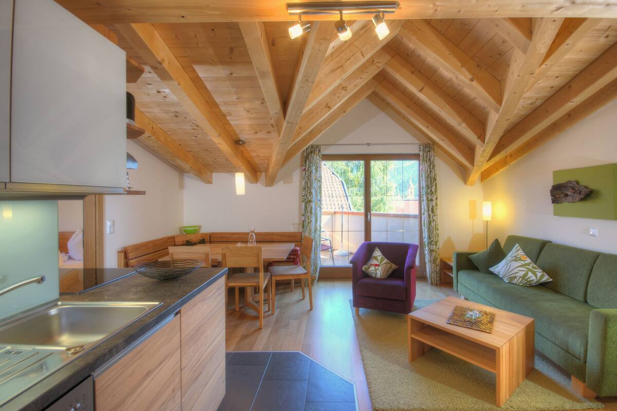 A modern kitchen with wooden beams, a dining area with a table and chairs, a green sofa, and a small coffee table.