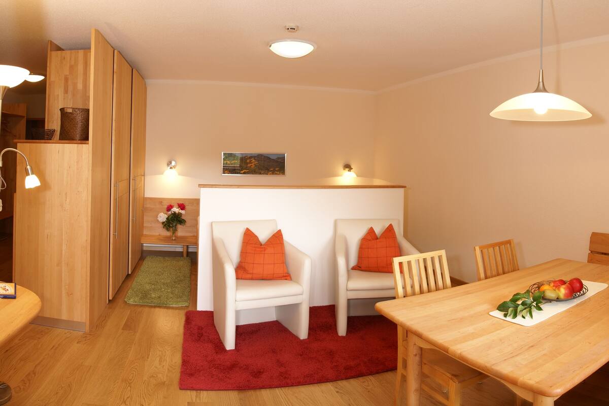 A cozy room with a wooden dining table, two white chairs with orange cushions, a red rug, and a wooden cabinet.
