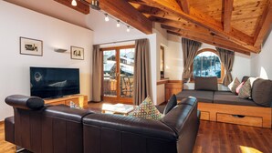 A modern living room with a leather sofa, a TV, wooden beams, and a large window with curtains.