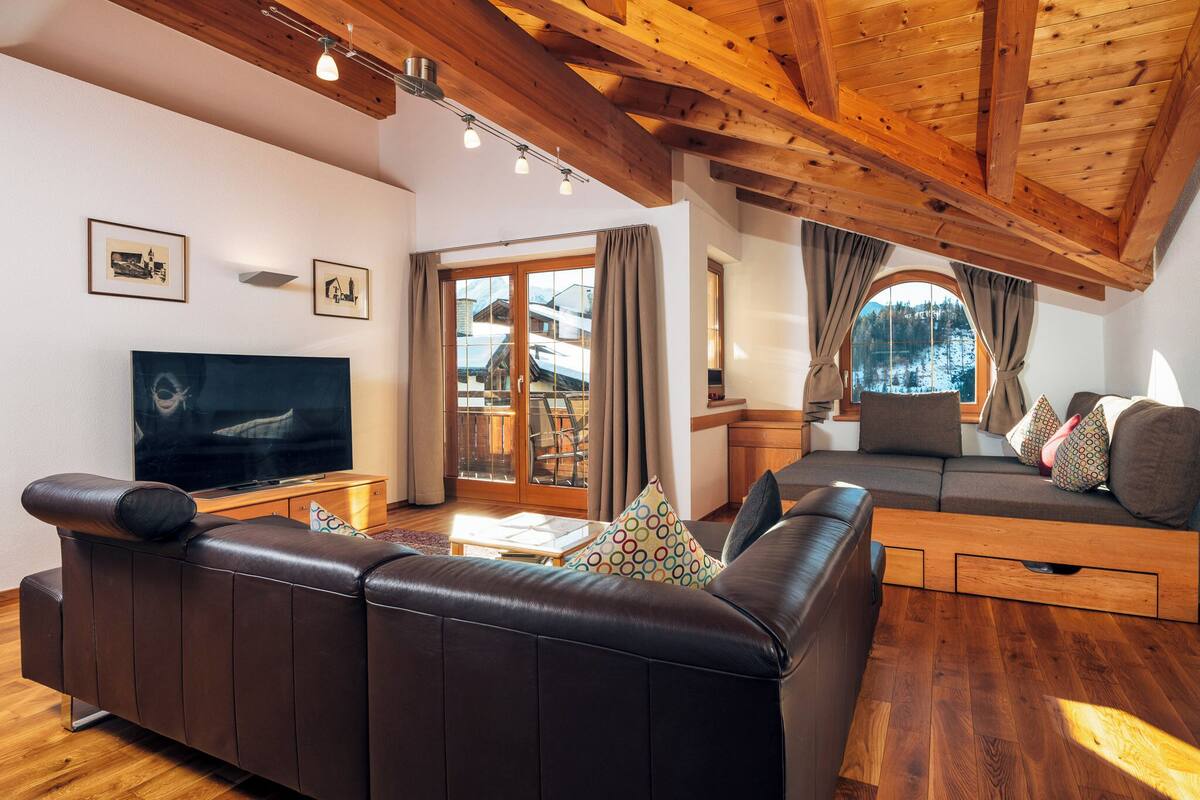 A modern living room with a leather sofa, a TV, wooden beams, and a large window with curtains.
