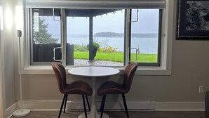Une pièce dotée d’une grande fenêtre donnant sur un plan d’eau et sur la verdure. On y trouve une table ronde avec deux chaises et un lampadaire blanc.