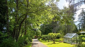 Un sentier dallé traverse un jardin luxuriant, agrémenté d’arbres et d’une serre.