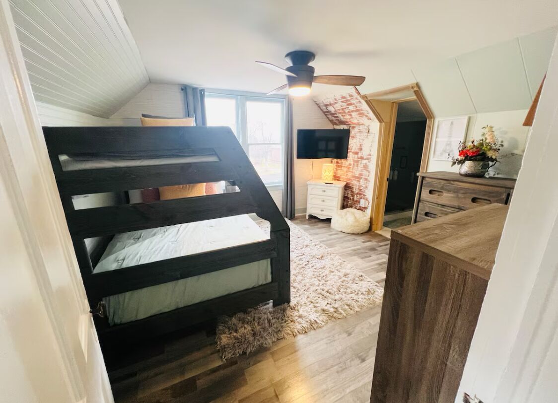 A bedroom with a bunk bed, a ceiling fan, a desk, a chair, a small rug, and a window with a view.