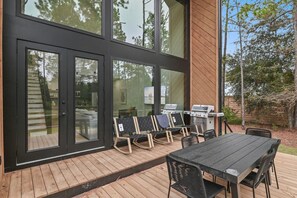 A modern outdoor patio with a wooden deck, black-framed glass doors, and a barbecue grill. - Modern Glass-Front Retreat l Stunning Nature Views (Santa Rosa Beach)
