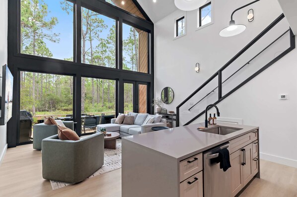 A modern interior with a kitchen, dining area, and seating space with large windows offering a forest view. - Modern Glass-Front Retreat l Stunning Nature Views (Santa Rosa Beach)