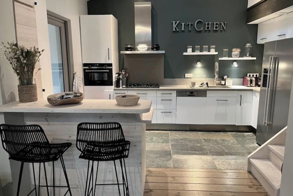 A modern kitchen with white cabinets, a stainless steel range hood, and a 'KITCHEN' sign on the wall. There are two black bar stools at the island.