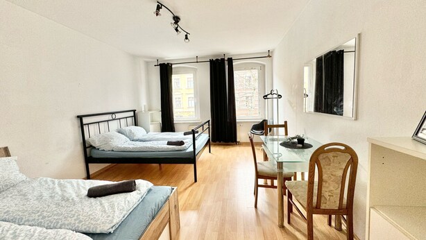 A compact room with a bed, a small table, and a chair. There is a window with curtains, a hanging light fixture, and a coat rack. - Modern apartment for sale in Chemnitz (Chemnitz)
